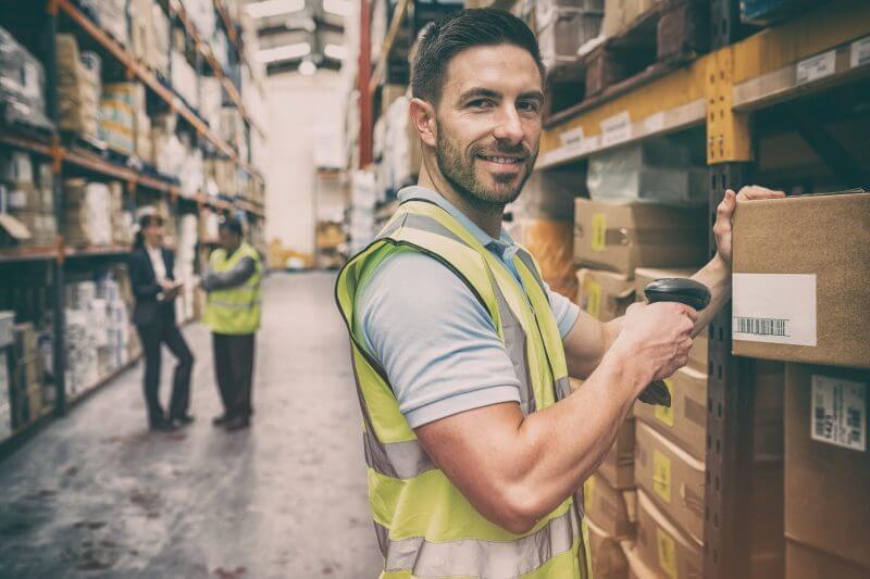 Smiling warehouse worker wearing a high-visibility vest, scanning a barcode on a cardboard box, with two colleagues in the background, engaged in conversation amongst shelves of products.