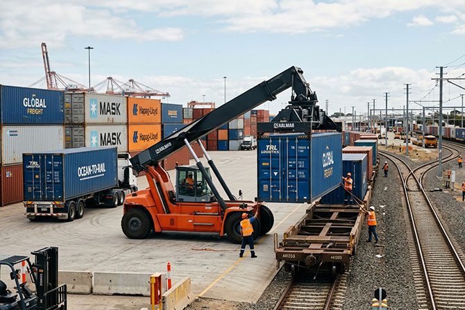 An orange reach stacker transferring a blue shipping container from a truck to a railcar at a busy logistics terminal.