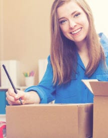 Smiling woman in a blue shirt at her desk, filling out a shipping label next to two cardboard boxes, with a computer in the background.