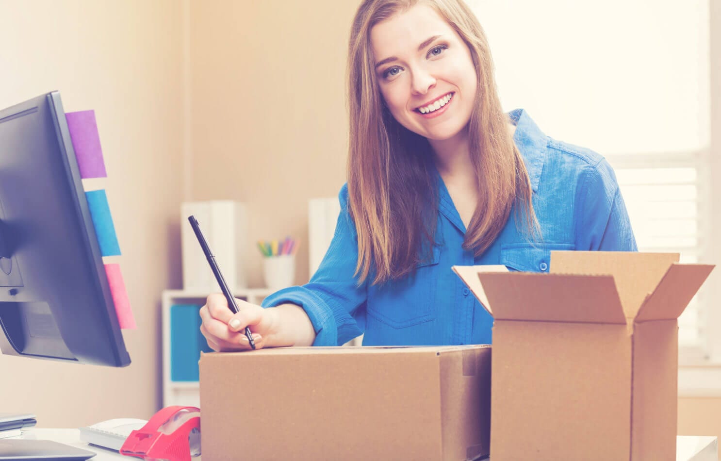 Smiling woman in a blue shirt at her desk, filling out a shipping label next to two cardboard boxes, with a computer in the background.