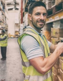 Smiling warehouse worker wearing a high-visibility vest, scanning a barcode on a cardboard box, with two colleagues in the background, engaged in conversation amongst shelves of products.