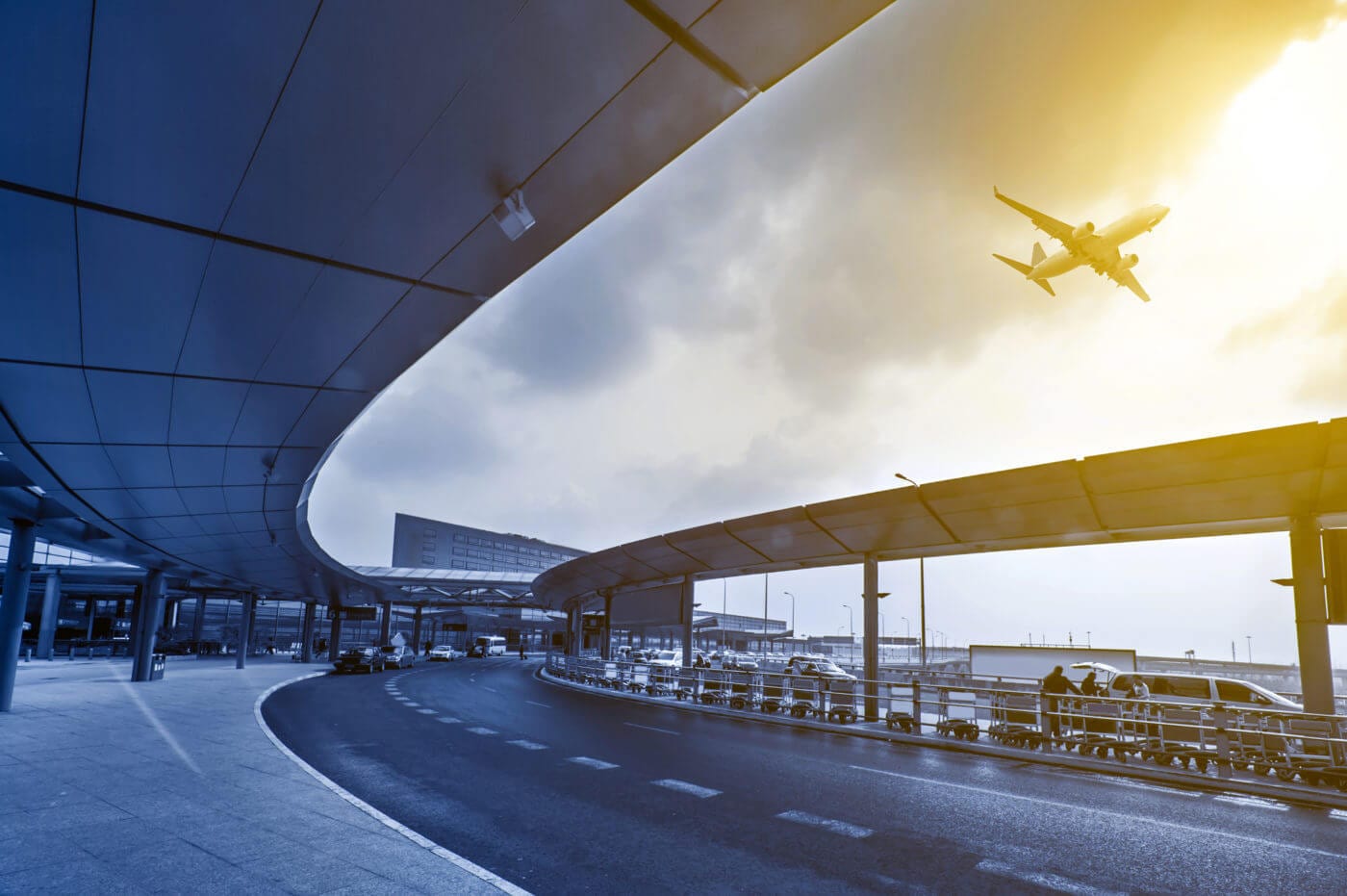 Scene of an airport terminal with a curved covered walkway, as an airplane approaches for landing against a backdrop of a cloudy sky and a warm, golden light.